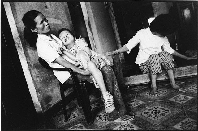A mother coddles her son, afflicted with Spina Bifida, while his blind sister sits close by.  The children's father fought in Cambodia just after the American conflict.